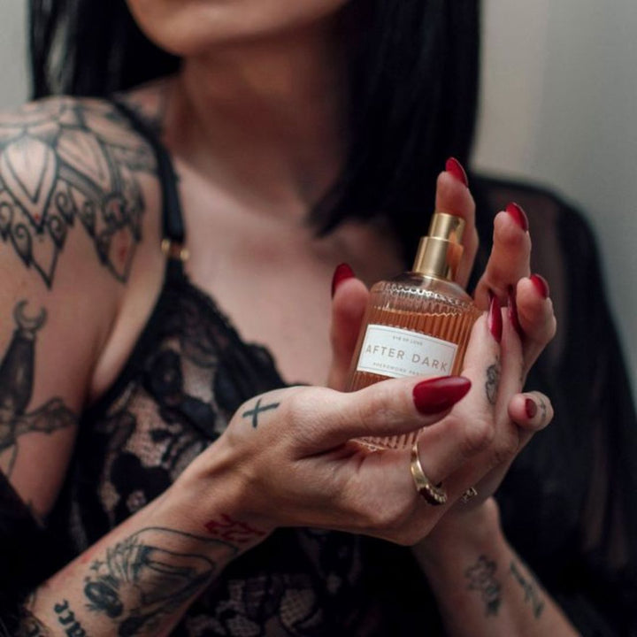 A tattooed woman with red-painted nails holding a bottle of After Dark pheromone perfume from Eye of Love.