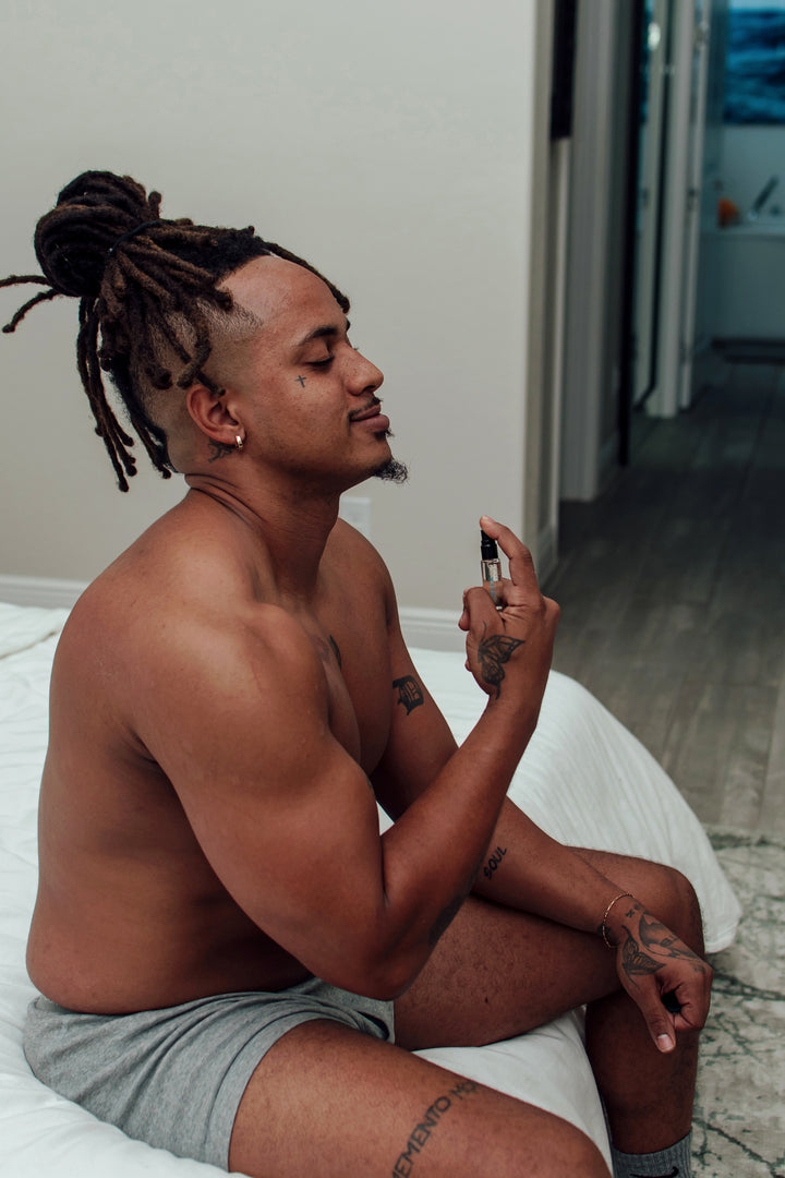 A smiling topless man with dreadlocks tied in a bun on a bed, applying Eye of Love travel-sized pheromone cologne.