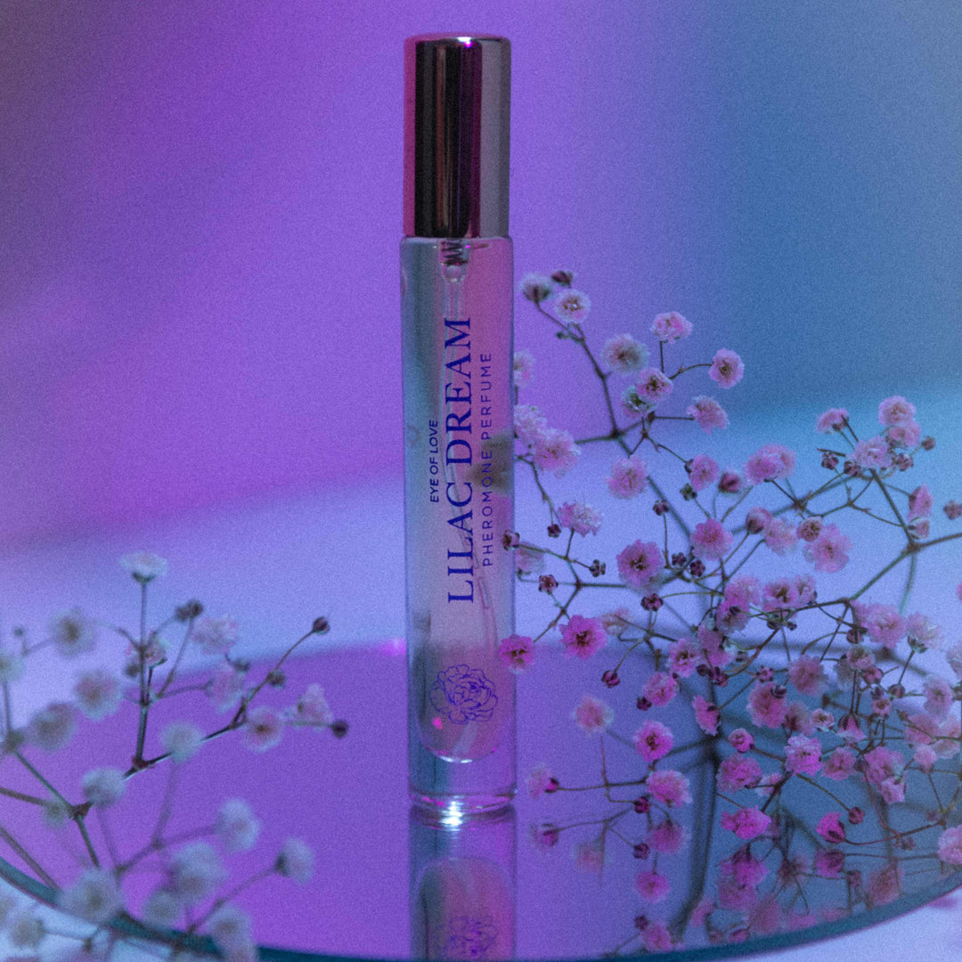 Lilac Dream Pheromone perfume on a mirrored circle, surrounded by sprigs of wildflowers and soft neon purple light effects.